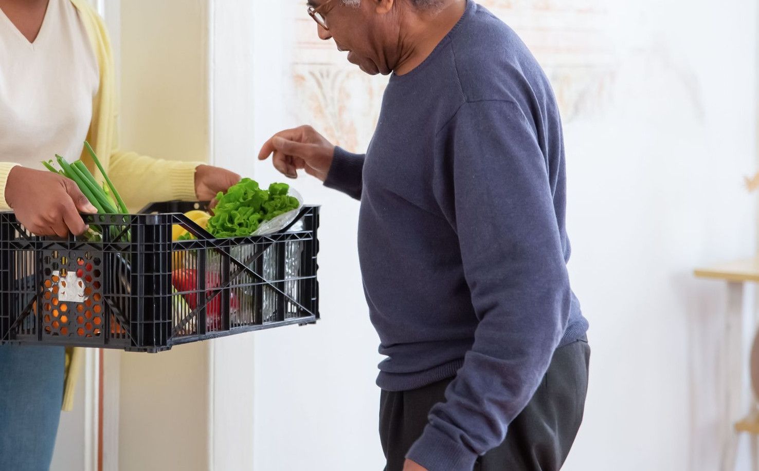 A young adult woman helps a senior man by carrying a crate of fresh vegetables indoors, symbolizing care and support.
